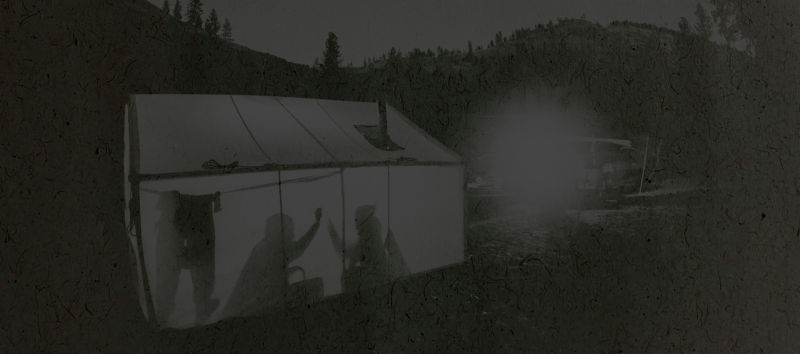 Black and white image of a campsite with two people silhouetted inside their tent.