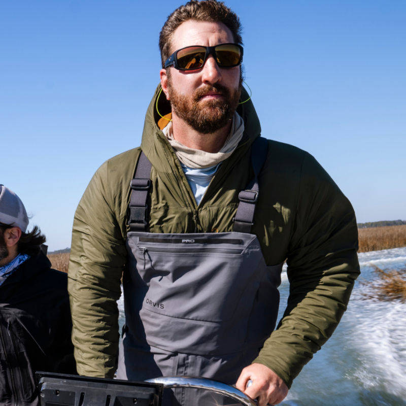 An angler piloting a speed boat while wearing an olive insulated jacket and waders.