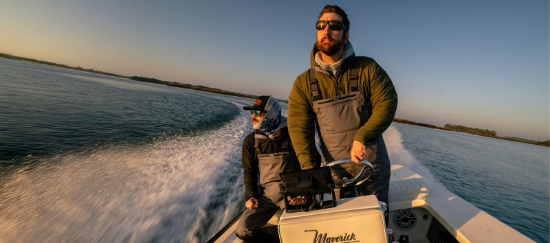 Two angler wearing waders on a speed boat facing into the sun.
