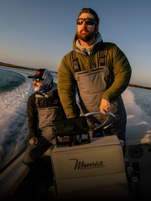 Two angler wearing waders on a speed boat facing into the sun.