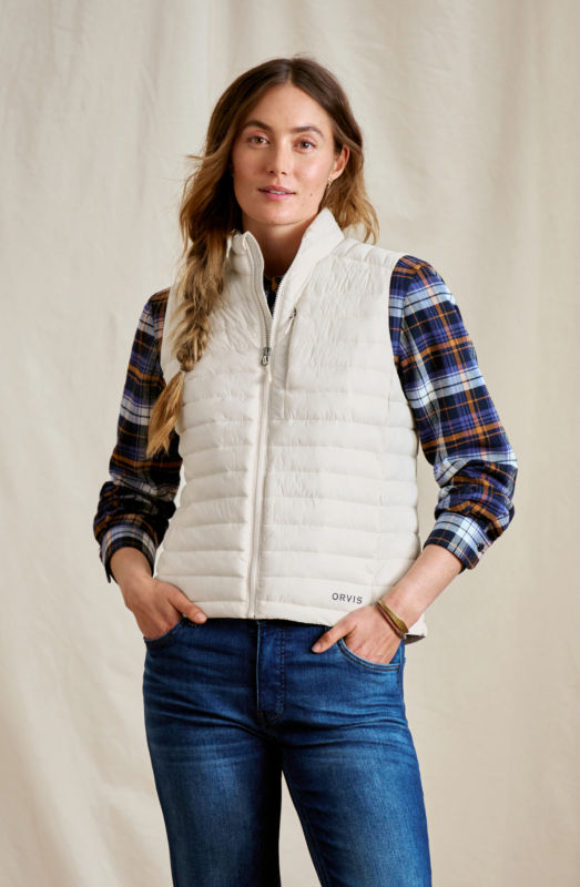 A woman wearing  a light cream vest over a blue and yellow flannel shirt with jeans.
