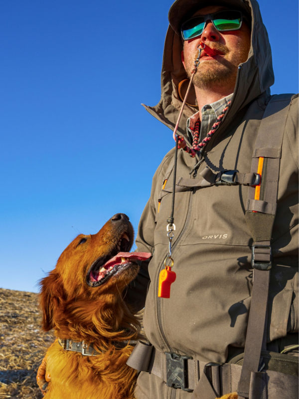 A hunter blowing a whistle with his dog by his side.