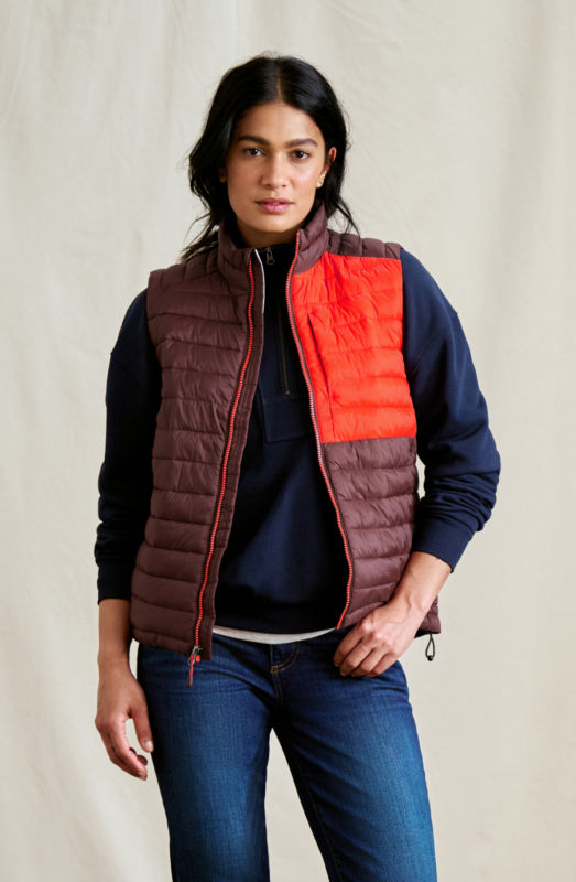 A model stands in front of a canvas curtain wearing a color-blocked vest over a navy sweatshirt and blue jeans.