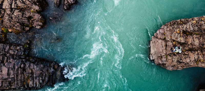 A drone view of a man standing on a rock fishing into swirling emerald white waters..