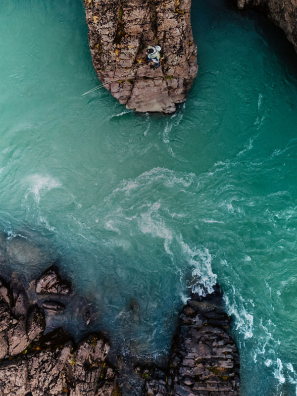 A drone view of a man standing on a rock fishing into swirling emerald white waters..
