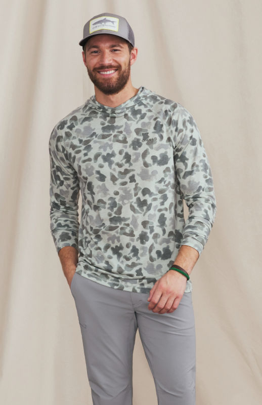 A man wearing a light green camo shirt over gray pants with a gray ball cap.