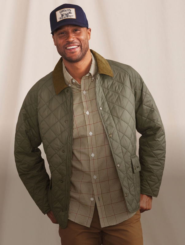 A smiling man stands before a cream curtain wearing a quilted Barbour jacket over a plaid button-down.
