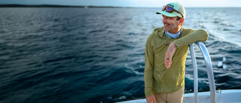 A man stands on a boat wearing an olive sun protective hoodie and sunglasses resting on his ball cap brim.
