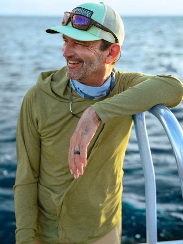A man stands on a boat wearing an olive sun protective hoodie and sunglasses resting on his ball cap brim.