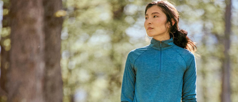 A woman stands outside under trees wearing a vivid blue sun protective quarter-zip.