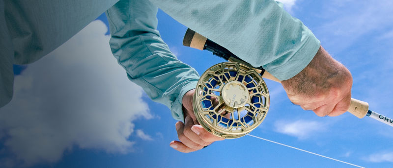 Two hands work a fly rod with a gold Ratio fly reel against the background of a bright blue sky.