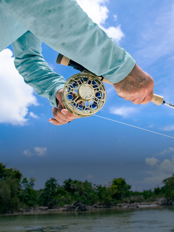 Two hands work a fly rod with a gold Ratio fly reel against the background of a bright blue sky.
