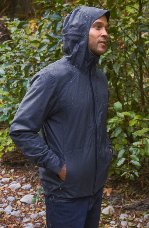 A man stands next to a bush wearing a navy raincoat with the hood up.