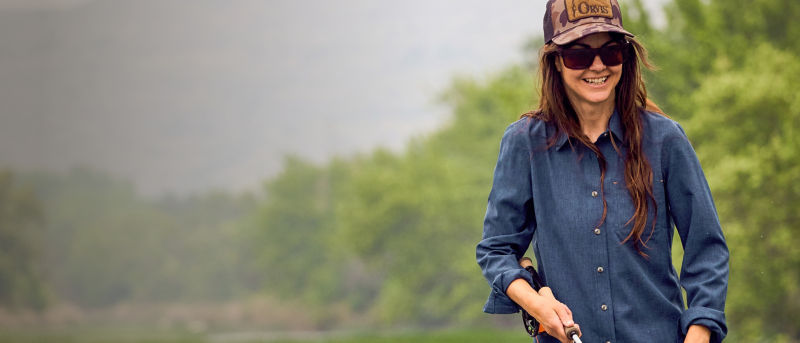 A woman in a blue tech chambray shirt and brown camo ball cap holding a fishing rod with lots of trees in the background.