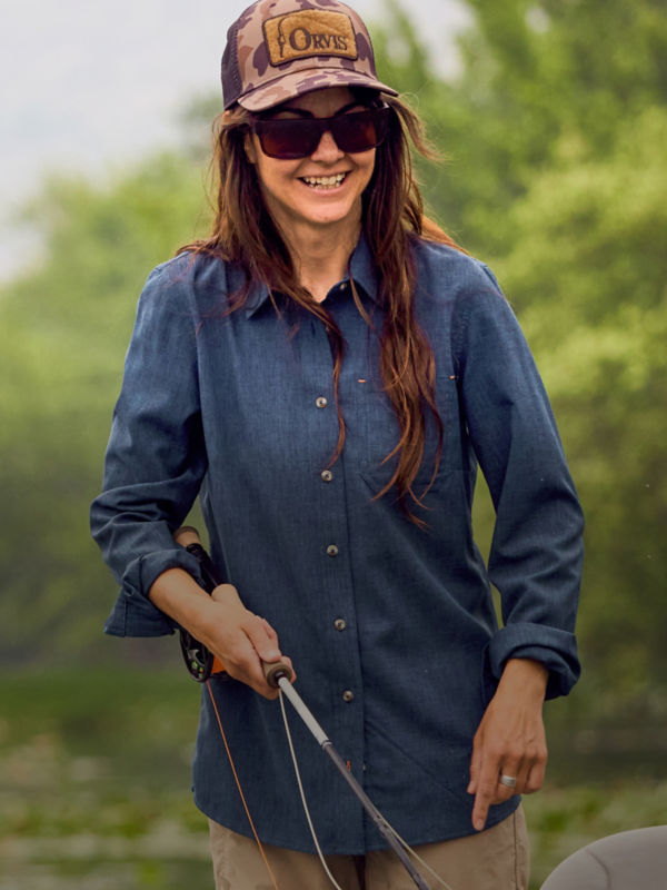 A woman in a blue tech chambray shirt and brown camo ball cap holding a fishing rod with lots of trees in the background.