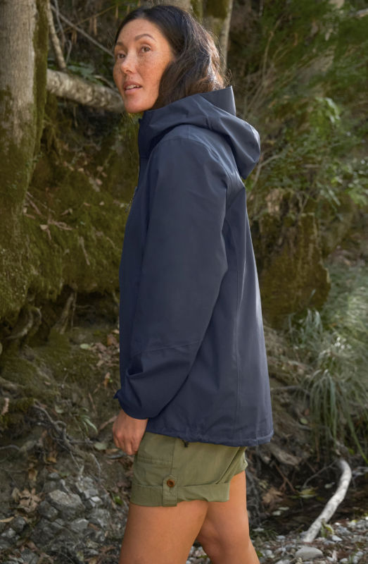 A woman wearing  a navy raincoat with olive shorts stands by a mossy bank.
