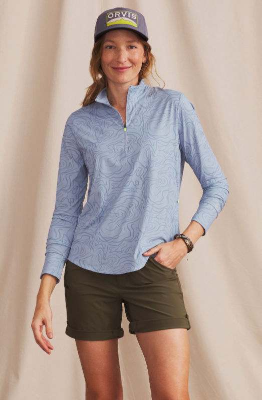 A woman is wearing a light blue printed top over olive shorts and a navy ball cap.