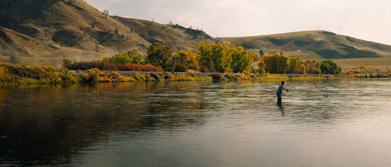 A gorgeous hilly landscape surrounding a wide river with an angler fishing.