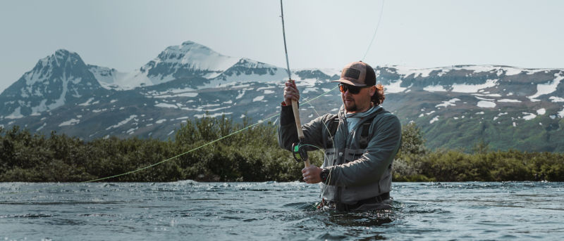 An angler in waders stands waist deep in a river casting under snowy peaks.