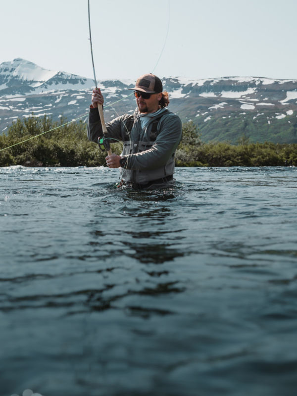 An angler in waders stands waist deep in a river casting under snowy peaks.