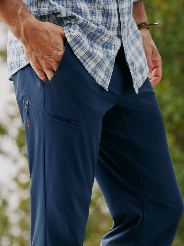 A man's hand in the pocket of navy technical pants with a blue and white plaid button-down.