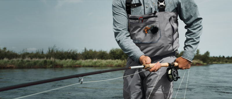 An angler in waders with a fly rod down by his hip.