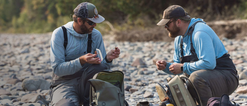 Two anglers wearing light blue hoodies and waders sitting on a rocky beach choosing flies from a fly box.
