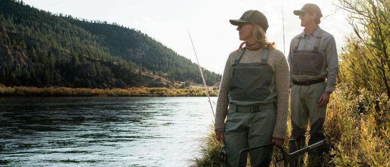 Two anglers wearing waders and carrying fly rods walk a path down to the river/