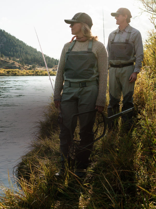 Two anglers wearing waders and carrying fly rods walk a path down to the river/