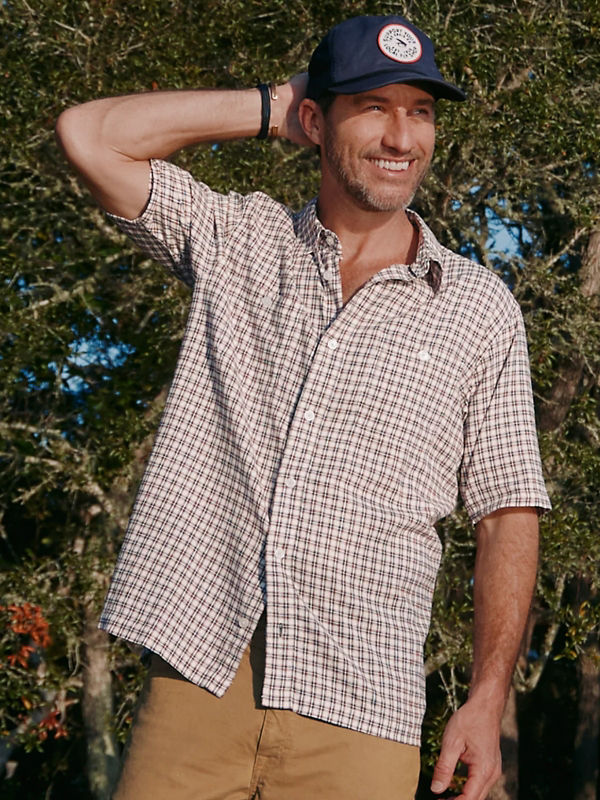 A model is wearing a short sleeve button down brown plaid shirt over tan shorts with a blue cap.