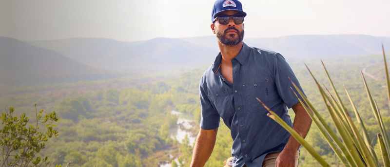 A man wearing a blue button-down shirt over tan pants and a blue hat hiking up a hill.