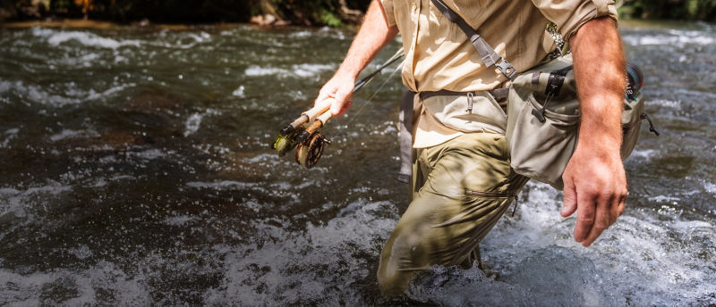 A man wading through whitewater carrying fishing gear.