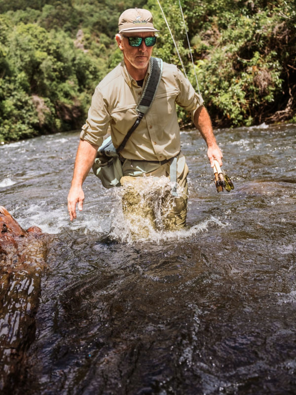 A man wading through whitewater carrying fishing gear.