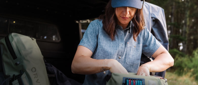 A woman wearing a blue chambray shirt and a blue hat looking into her fishing pack.