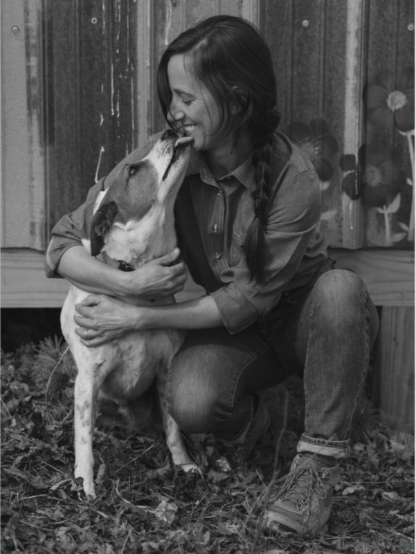 A woman squats down to hug a dog who licks her face.