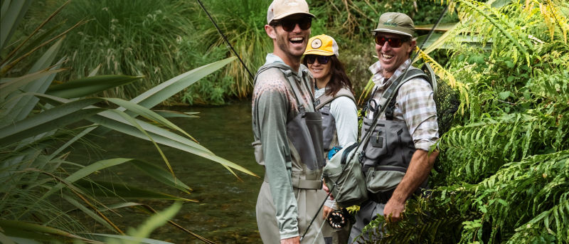 Three anglers in fishing gear turn with smiles from the river.
