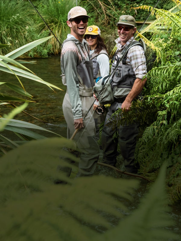 Three anglers in fishing gear turn with smiles from the river.