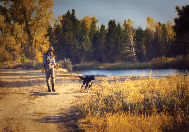 A woman walking wearing waders while her dog runs by her side.