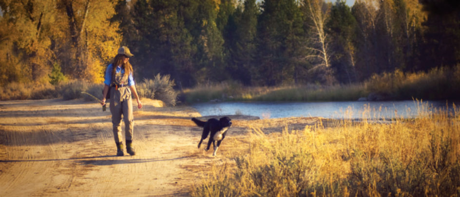 A woman walking wearing waders while her dog runs by her side.