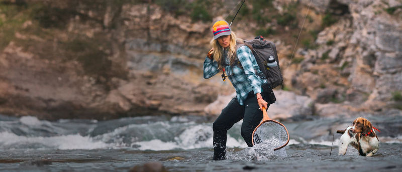 A woman wearing a blue plaid shirt and a bright cap is fishing with her dog.