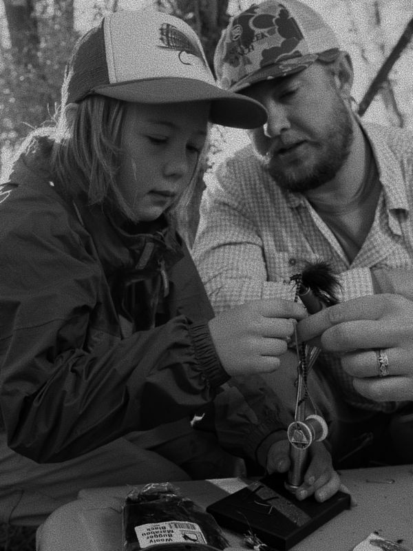 A man shows a fly on a fly-tying vise to a child.