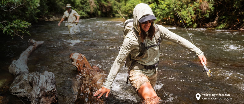 Two anglers loaded down with gear wade through a fast-moving river. In the bottom corner a location tag with the words: Taupo, New Zealand, Chris Jolly Outdoors.