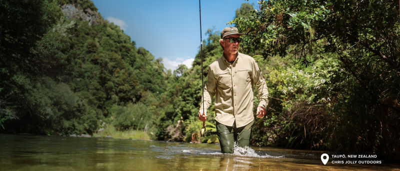 An angler in khaki wades knee deep through a slow moving river.