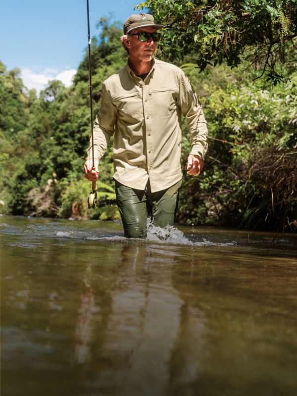 An angler in khaki wades knee deep through a slow moving river.