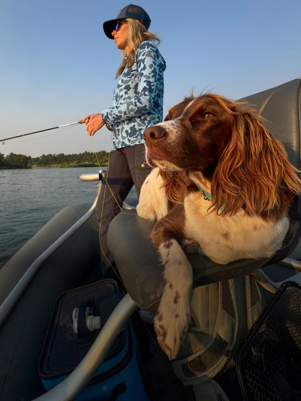 An angler fly fishes from the back of a boat while her dog lays on a boat chair.