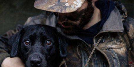 A hunter in full camo embraces a young black Labrador Retriever.
