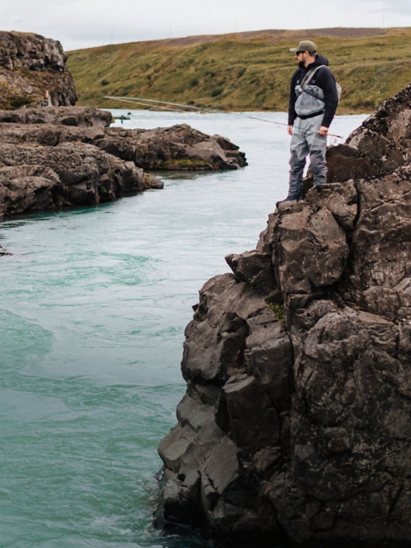 An angler stands atop a steep rock bank looking down into a crystal blue river.