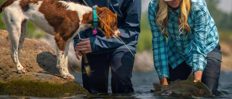 Two anglers with a just-caught fish in hand and an eager spaniel looking on.