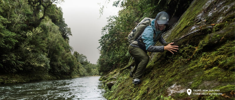 An angler wearing a waterproof backpack climbs on moss covered rocks at the river's edge wearing Jackson Quick-Dry Pants.
