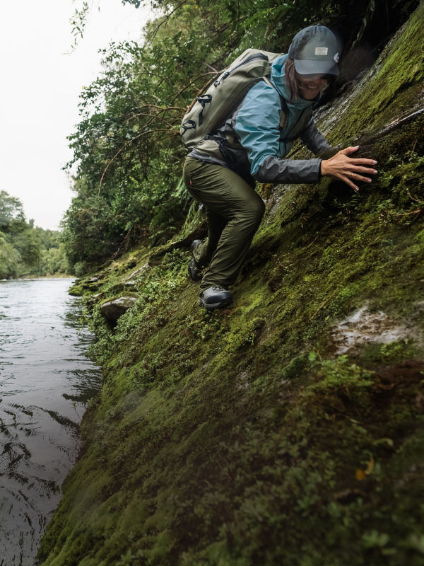 An angler wearing a waterproof backpack climbs on moss covered rocks at the river's edge wearing Jackson Quick-Dry Pants.